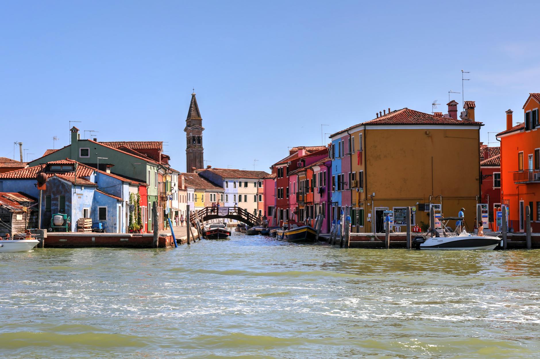 Colorful houses lining a canal with boats docked and a bell tower in the background under a clear blue sky