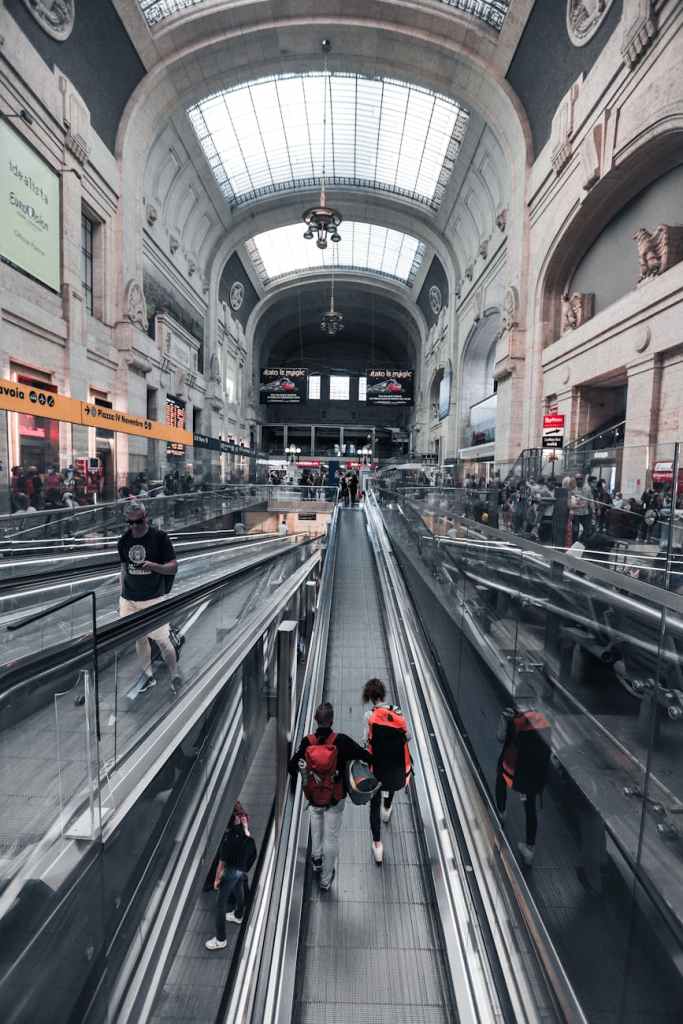 Interior view of a busy train station with high ceilings, showing escalators and travelers moving through the space.