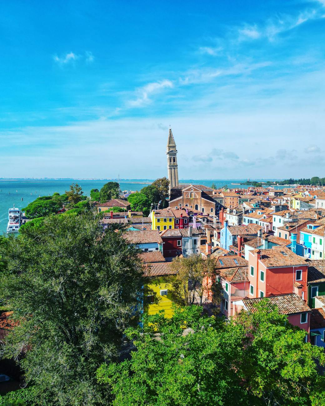 Colorful houses with terracotta roofs near green trees and a tall leaning bell tower by a large body of water under a blue sky