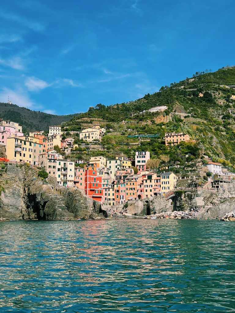Colorful coastal houses on a hillside in Cinque Terre, Italy, with a clear blue sky and calm waters in the foreground.