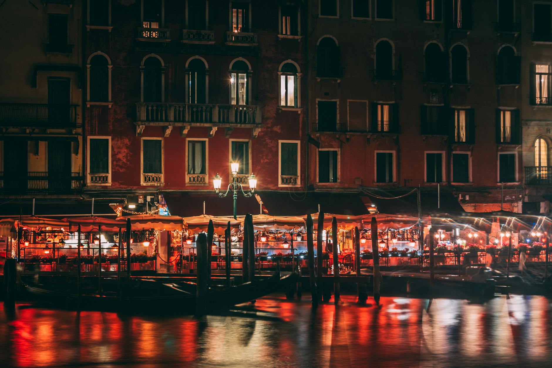 Nighttime view of a canal-side building with illuminated windows, lit street lamps, and outdoor seating areas reflected in the water