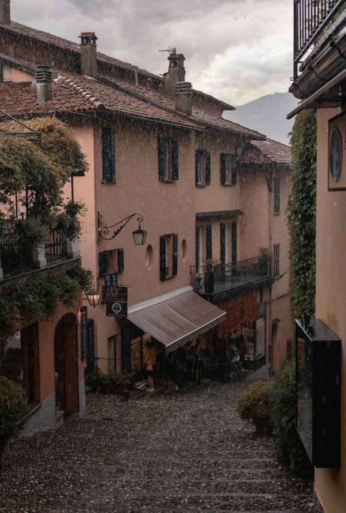 A picturesque cobblestone street in a quaint town, with weathered buildings featuring shutters and awnings, under heavy rainfall.