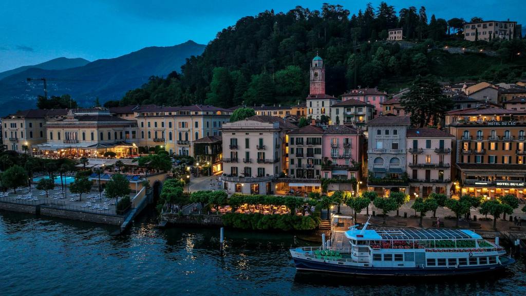 A scenic view of a lakeside town at dusk, featuring charming buildings, a marina with a boat, and lush green hills in the background.