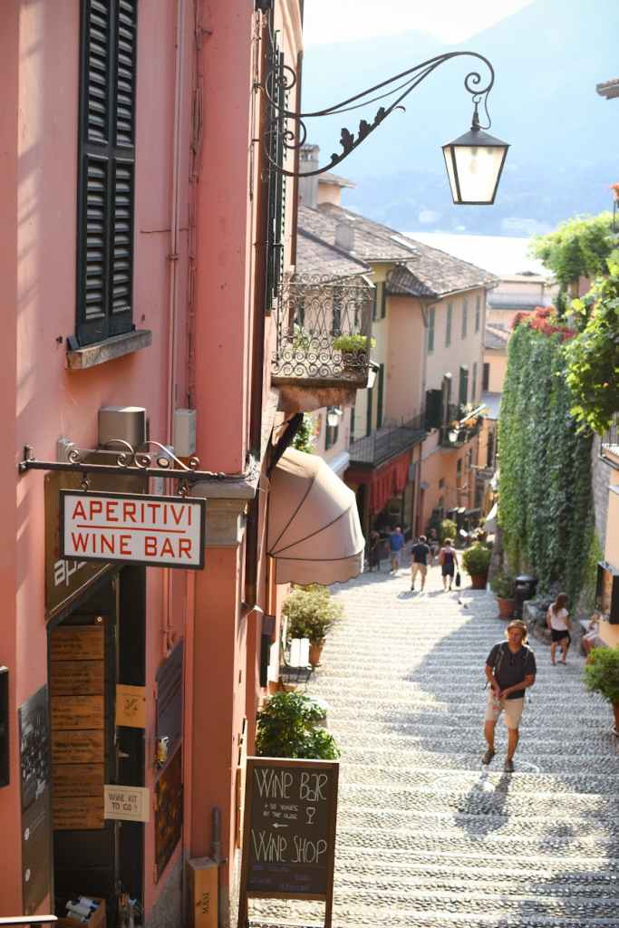 A picturesque street scene in a quaint town featuring a 'Aperitivi Wine Bar' sign, cobblestone pathway, and lush greenery. People stroll leisurely, enjoying the charming atmosphere.