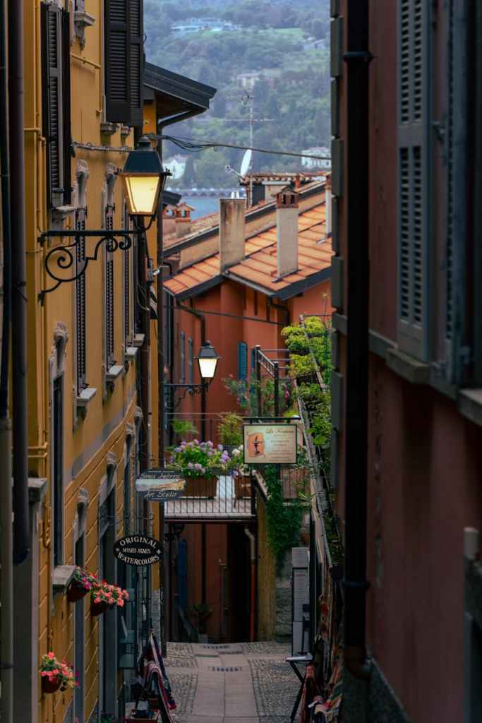 A narrow, cobblestone alleyway lined with colorful buildings, hanging flower pots, and a street lamp, leading to a quaint entrance with signs indicating local shops.