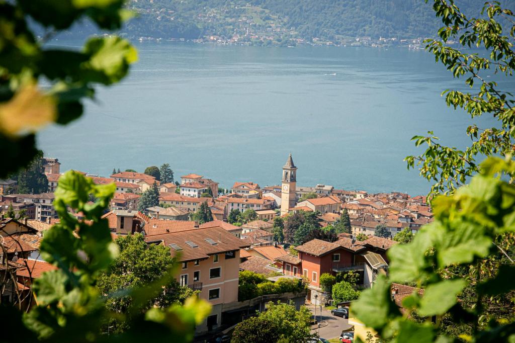 A scenic view of a lakeside village with terracotta-roofed buildings, a clock tower, and lush greenery in the foreground, overlooking a calm lake surrounded by hills.