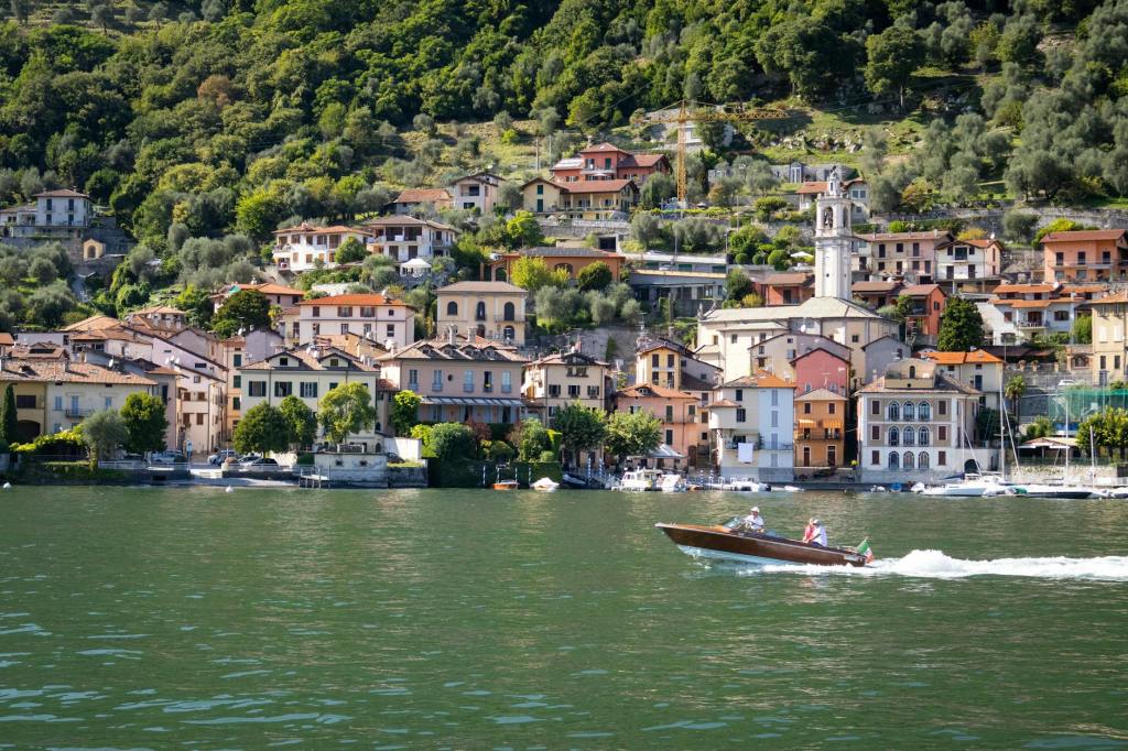 A scenic view of colorful houses along the shoreline of a lake, with a boat speeding across the water and hills in the background.