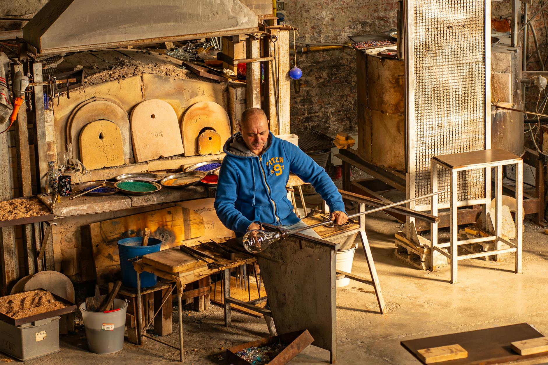 Man shaping molten glass in a workshop with a furnace and tools