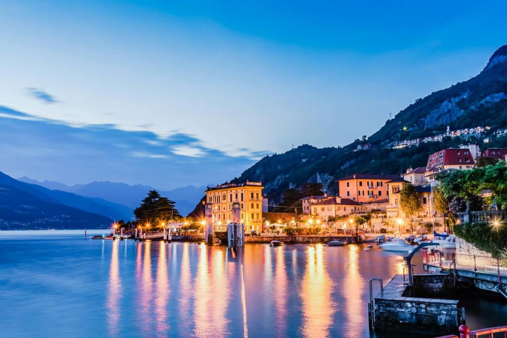 Scenic view of a lakeside town at dusk, featuring illuminated buildings reflected in the calm water, surrounded by mountains.