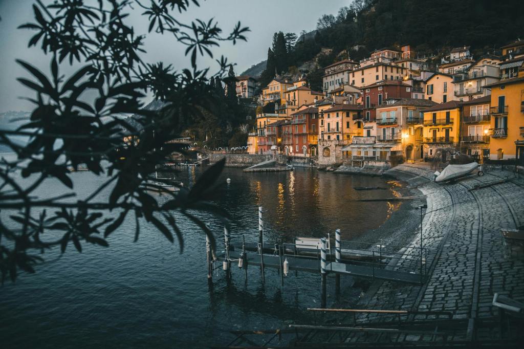 A serene lakeside view with colorful buildings along the shore, illuminated by evening lights, framed by leafy branches in the foreground.