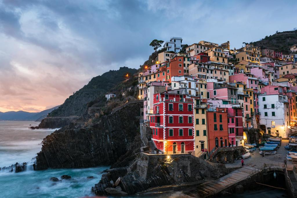 Colorful buildings on a rocky coastline during sunset, with a calm sea and mountains in the background.