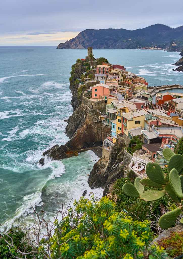 A picturesque coastal village perched on a rocky cliff, overlooking turbulent waves and surrounded by lush greenery and mountains in the background.