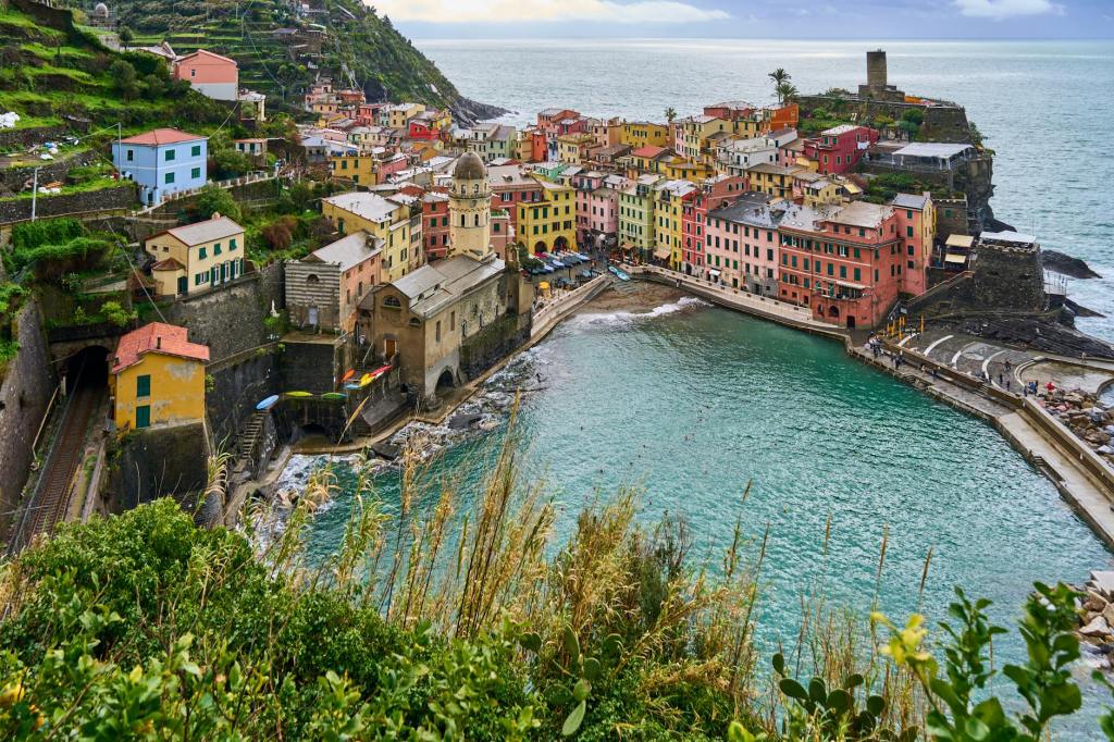 A panoramic view of a colorful coastal village with buildings in various hues overlooking a tranquil bay, surrounded by green hills and rocky cliffs.
