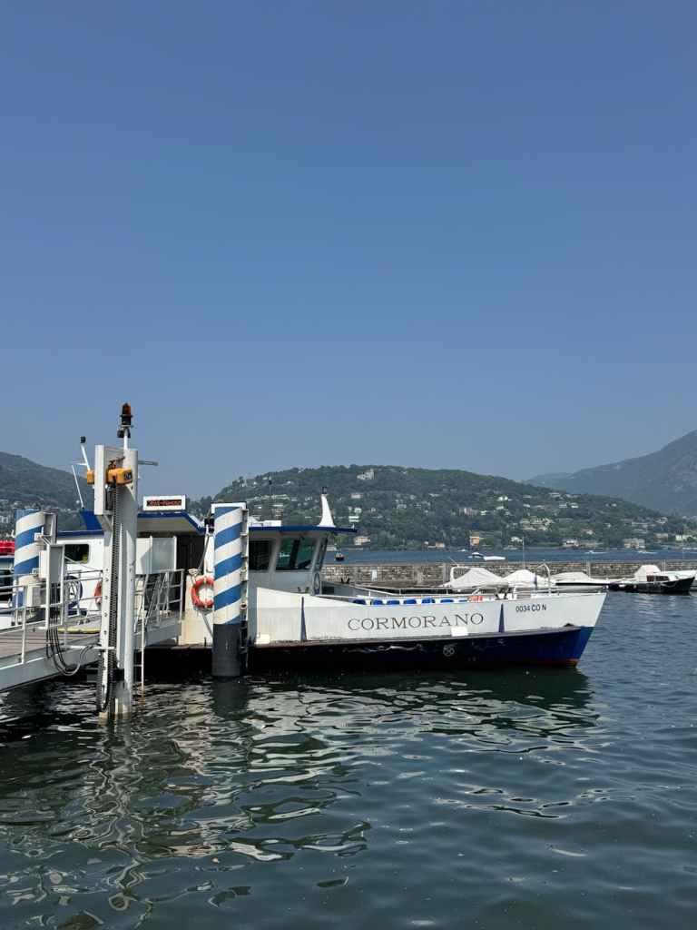 A boat named 'CORMORANO' docked at a pier on a calm body of water, surrounded by green hills and clear blue skies.