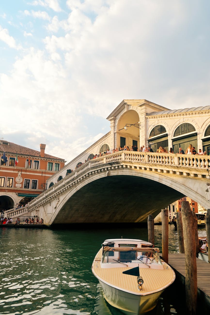 People walking and standing on the Rialto Bridge over the Grand Canal in Venice with a boat docked in the foreground