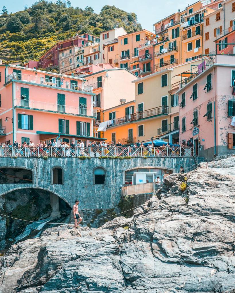 A coastal scene featuring colorful buildings stacked on a hillside, overlooking a rocky shore. A shirtless man stands on the rocks in the foreground, while a crowd gathers on a balcony above.
