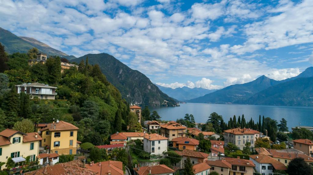 A scenic view of a lakeside village surrounded by mountains, featuring colorful homes with terracotta rooftops and lush greenery against a backdrop of blue skies and scattered clouds.