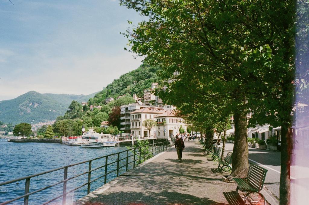 Scenic lakeside walkway with trees, benches, and a person strolling, overlooking a lake surrounded by mountains and buildings.