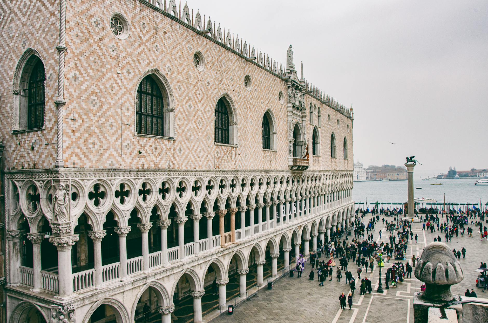 The Doge's Palace in Venice with an ornate facade, arched windows, statues, a plaza filled with people, and a column topped by a lion statue by the waterfront