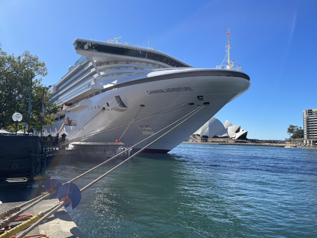 A cruise ship named 'Carnival Adventure' docked at Circular Quay in Sydney, with the Sydney Opera House visible in the background under a clear blue sky.