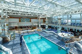 An indoor swimming pool on a cruise ship, featuring a transparent roof and several deck chairs around the pool area.
