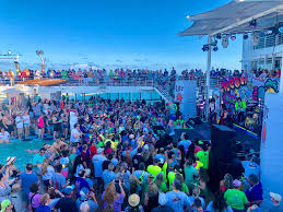 A lively crowd in 1980s-themed outfits gathers around a pool deck for a sail away party, with colorful decorations and a clear blue sky in the background.