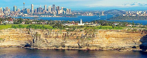 A panoramic view of Sydney, showcasing the skyline with tall buildings, the Sydney Harbour, and the iconic Sydney Harbour Bridge, set against a clear blue sky.