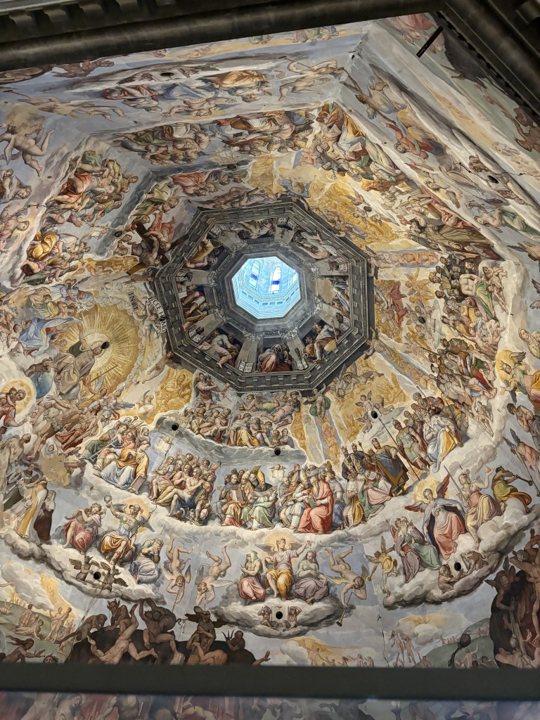 A view from below of the intricate frescoed ceiling of a dome, featuring religious scenes, angels, and figures surrounded by clouds, with a central skylight casting light into the space.