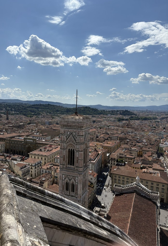 A panoramic view of Florence, Italy, showcasing the cityscape with terracotta rooftops, the prominent Bell Tower of Santa Maria del Fiore, and a bright blue sky dotted with clouds.