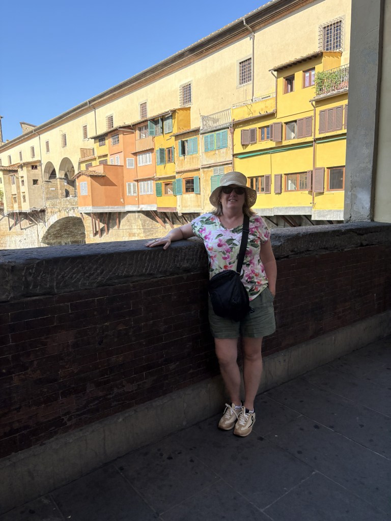 The author stands against a stone wall with the colorful buildings of the Ponte Vecchio in Florence, Italy, in the background. She is wearing a floral shirt, shorts, and a sun hat.