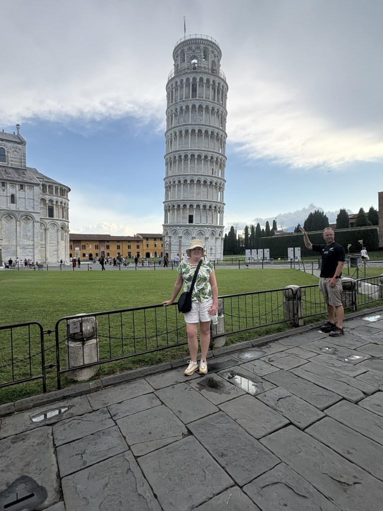 The author poses in front of the Leaning Tower of Pisa, surrounded by grass and historic buildings, with a cloudy sky in the background.