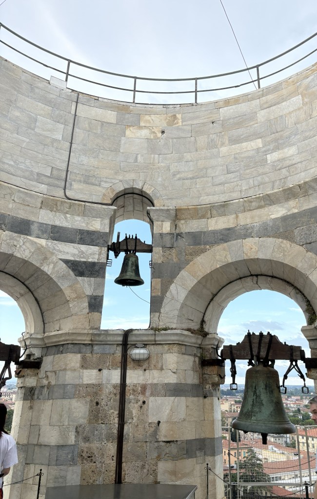 Interior view of a stone bell tower featuring two large bells hanging from arches, with a cityscape visible in the background.