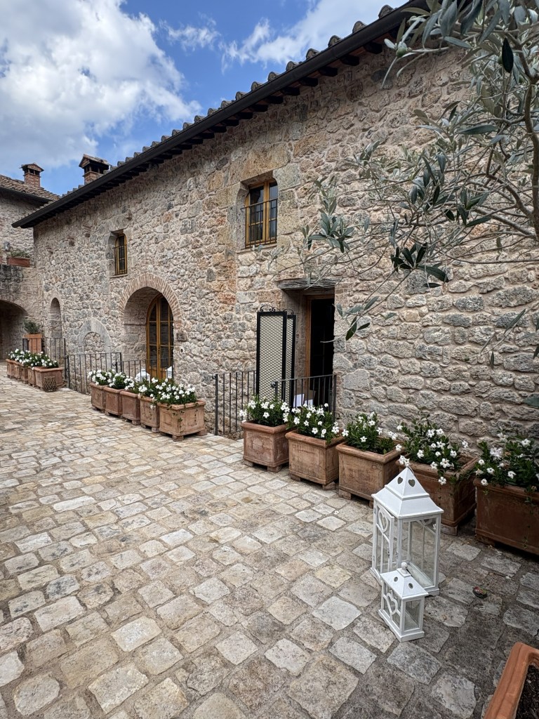A rustic stone building with windows and a courtyard filled with flower pots, beside a white lantern on a cobblestone path under a partly cloudy sky.
