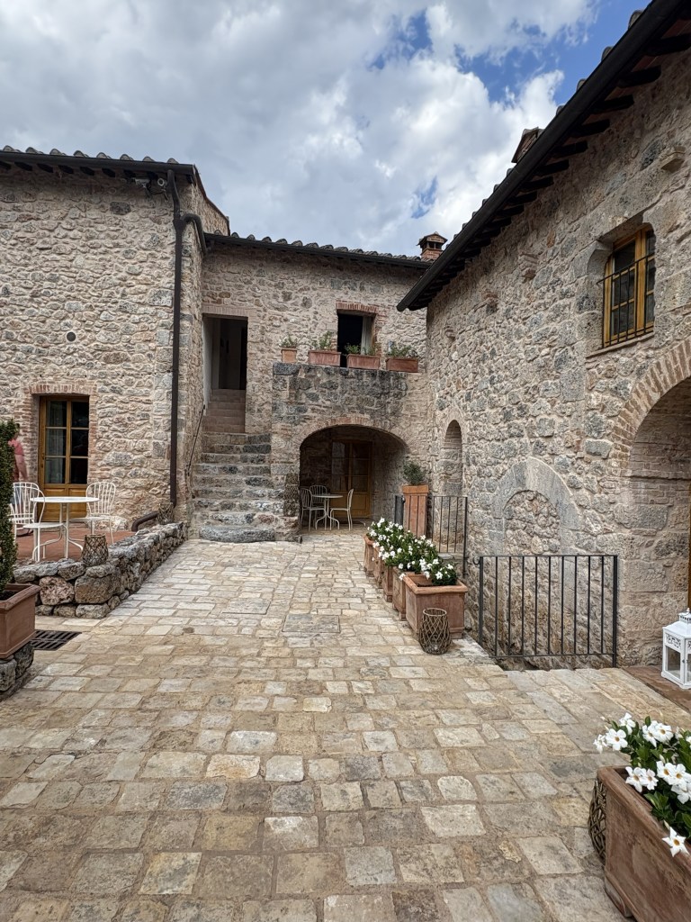 Stone courtyard of a rustic building, featuring potted flowers, light wooden furniture, and a staircase leading to an upper level under a cloudy sky.
