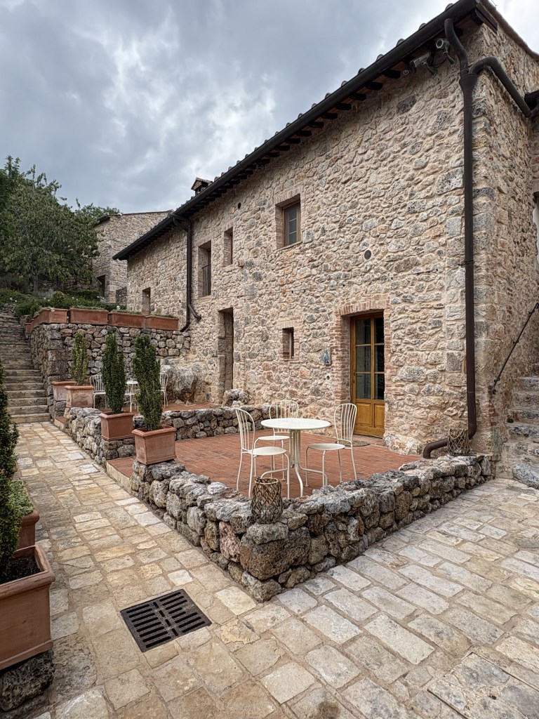 Exterior view of a rustic stone building with a terrace, featuring a small table and chairs, surrounded by potted plants and stone pathways under a cloudy sky.