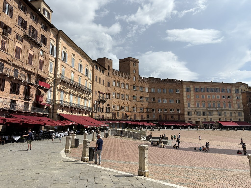 A view of a historic square in Siena, Italy, showcasing elegant buildings and outdoor dining under red awnings, with people strolling and sitting on the cobblestones.