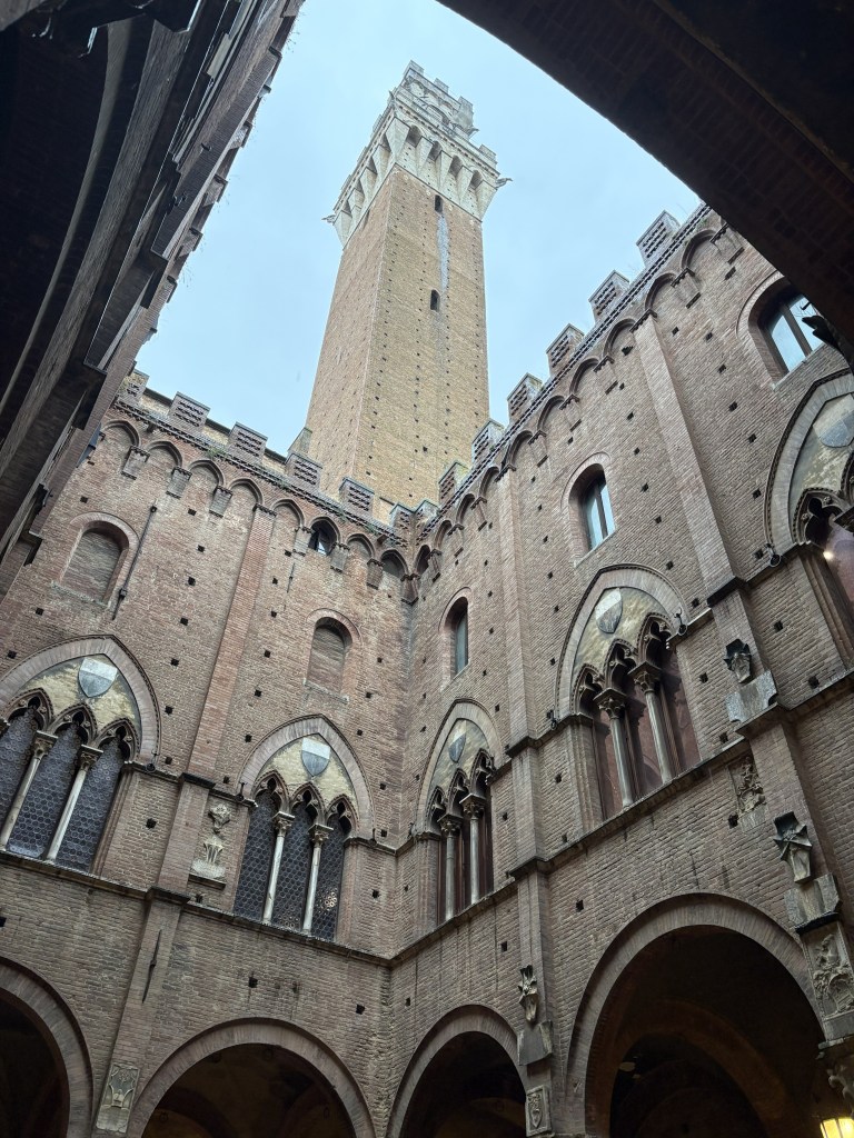 A view looking up towards the tall brick bell tower of the Palazzo Pubblico in Siena, Italy, framed by the archways of the courtyard with ornate windows and architectural details.