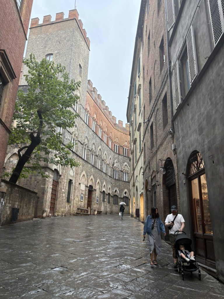 A narrow cobblestone street lined with historic buildings, featuring a tall tower in the background. Two people stroll down the wet pavement, one pushing a stroller, while another person walks by holding an umbrella.