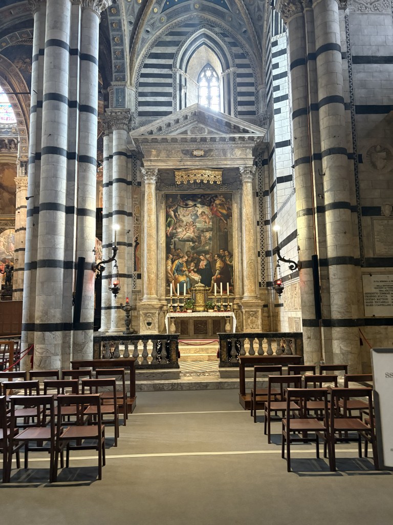 Interior view of a cathedral featuring tall, striped columns, an altar with a large painting, and wooden chairs arranged in front.