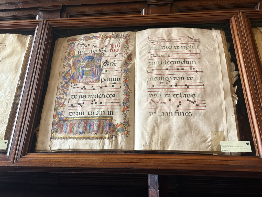 Open pages of an illuminated medieval manuscript displaying musical notation and decorative illustrations, housed in a glass display case.