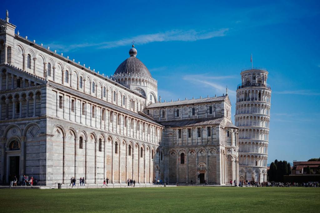 A view of the Leaning Tower of Pisa alongside historical buildings, with a clear blue sky in the background.