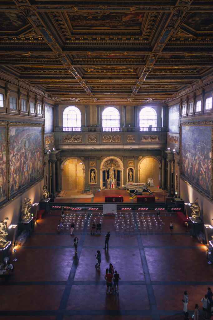 An interior view of a grand hall with ornate ceilings, featuring large murals on the walls, sculptures, and people sitting and walking around.