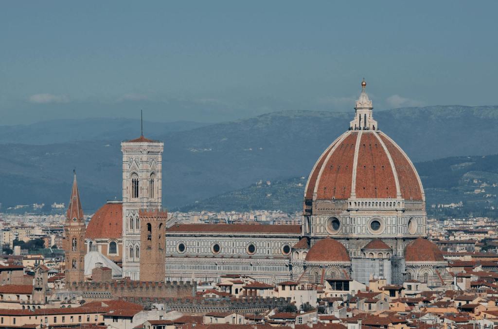 Panoramic view of Florence, Italy, showcasing the iconic dome of the Cathedral of Santa Maria del Fiore and the Giotto's Campanile tower, surrounded by historical buildings and a mountainous backdrop.