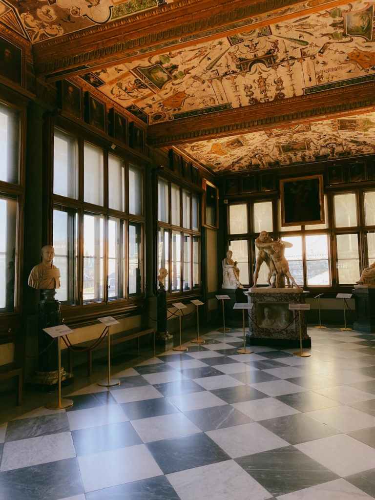 A beautifully decorated art gallery featuring a colorful frescoed ceiling, marble floor with checkerboard pattern, and several classical sculptures displayed along the walls.