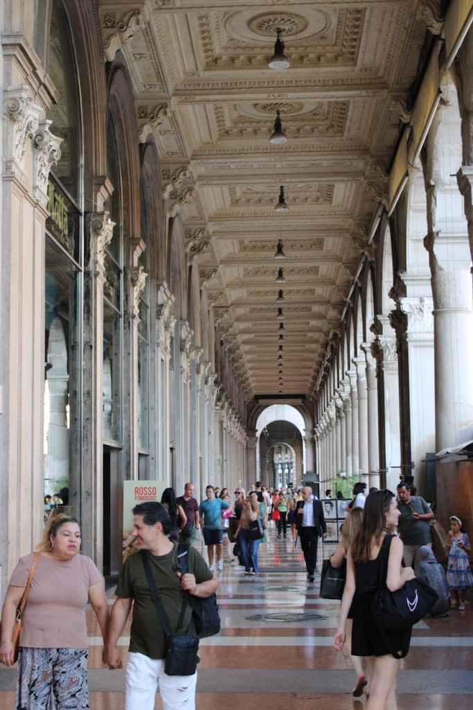A bustling corridor with ornate architecture featuring arches, columns, and intricate ceiling details. People stroll along the walkway, some chatting while others carry bags.
