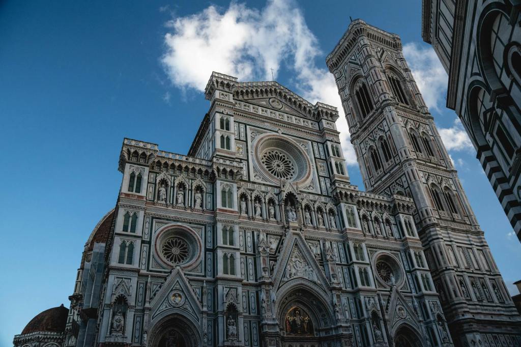 Facade of the Florence Cathedral featuring intricate designs and a tall bell tower, under a clear blue sky with clouds.