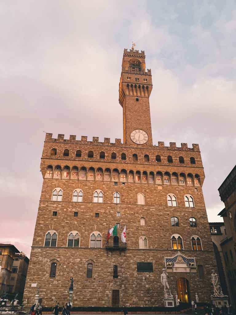 A tall medieval tower with a clock, part of a historic stone building in Florence, Italy, surrounded by figures and flags.