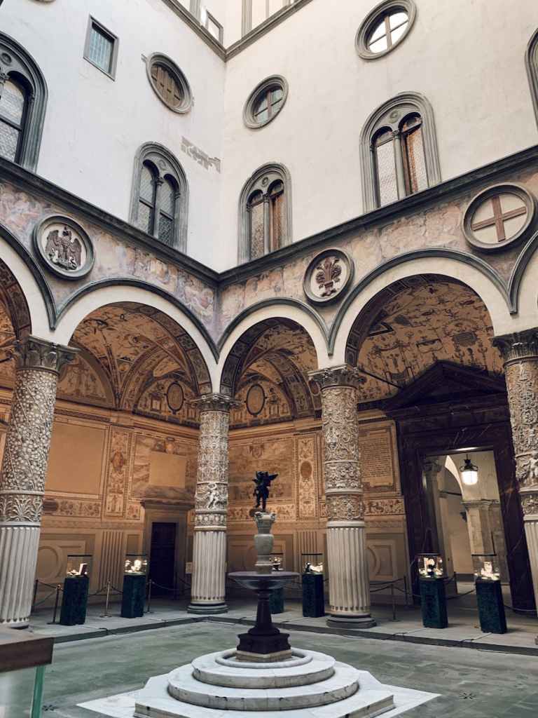 Interior courtyard of a historic building featuring decorative arches, detailed columns, frescoed walls, and a central fountain with a small statue.