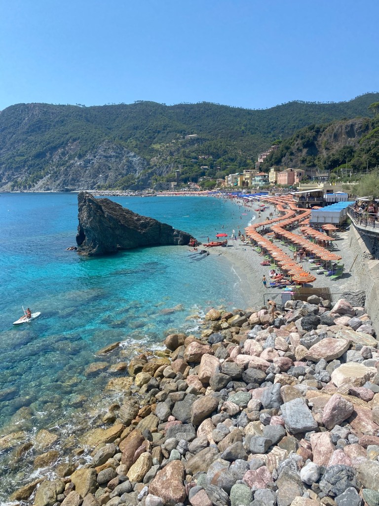 A scenic beach view featuring clear turquoise water, rocky shoreline, and colorful beach umbrellas lined along the shore with distant hills in the background.