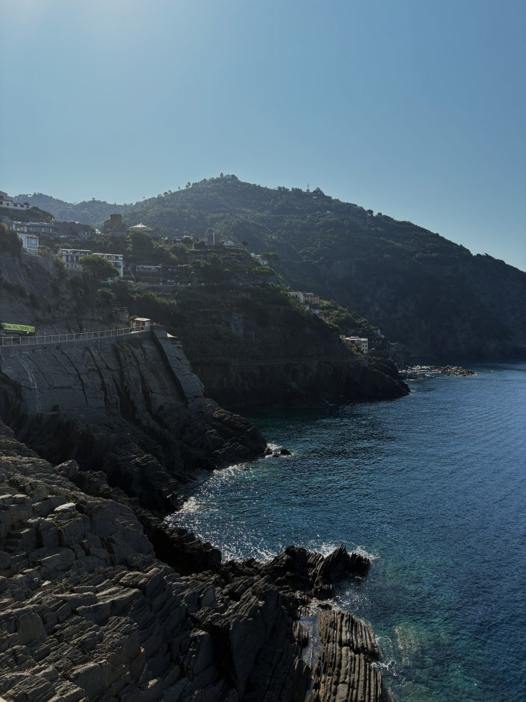 Scenic coastal view featuring rocky cliffs, clear blue water, and hills in the background under a bright sky.